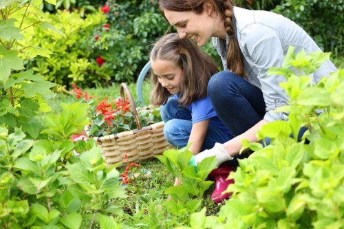 A woman and two children gardening outdoors in a lush, well-maintained garden with dense green foliage, flowering shrubs, and a clear grassy lawn in the foreground. The woman appears to be helping the children, who are crouched down and engaged in planting or tending to plants, with a wicker basket nearby. The garden features a variety of leafy plants, some with red flowers, and is bordered by a mixture of bushes and trees in the background. The scene is set on a sunny day with natural light illuminating the vibrant green leaves and colorful blossoms, creating a peaceful outdoor environment typical of a well-kept garden space in Belmont or nearby areas. The image emphasizes outdoor family gardening activities, with a focus on natural plant textures and garden layout suitable for professional gardening services like hedge trimming, lawn care, and general outdoor maintenance offered by Hedge Trimming Belmont.