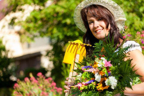 A smiling woman wearing a wide-brimmed straw hat stands outdoors in a garden, holding a bright yellow garden rake and a large bouquet of colorful flowers and greenery. Behind her, there is a lush garden scene with a well-maintained lawn, vibrant flower beds, and green foliage, suggesting a landscaped yard in Belmont. The garden features a mix of grass, flowering plants, and shrubbery, with some paved or gravel areas visible in the background. Natural sunlight illuminates the scene, highlighting the vibrant colors of the flowers and the greenery, creating a cheerful and inviting outdoor environment that reflects professional gardening and landscaping care provided by companies like Hedge Trimming Belmont.